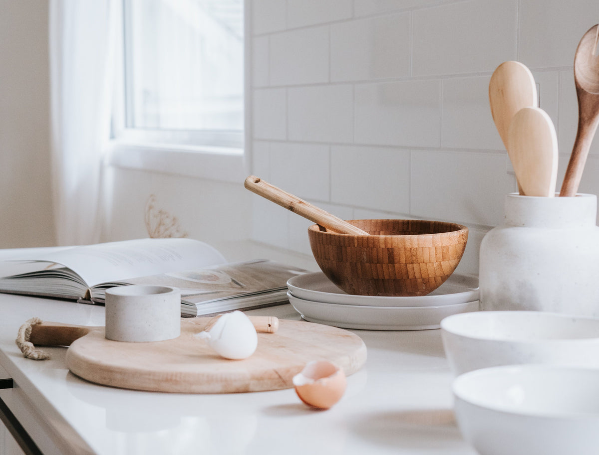 A styled kitchen countertop scene with a wooden bowl and wooden spoon resting inside a white ceramic utensil holder, alongside white bowls, a small wooden cutting board, eggs, and an open cookbook near a window.